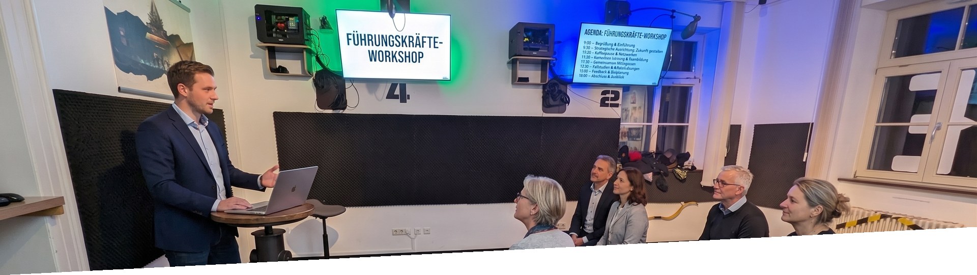 Leadership team in a workshop: consultant at a lectern, participants in a semicircle, screen titled leadership workshop